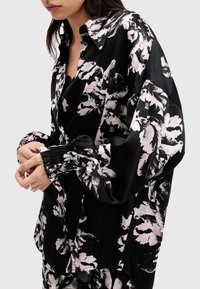Camisa negra con estampado floral en material satinado, de corte holgado, mangas largas y puños fruncidos, con grandes patrones florales en blanco y rosa.