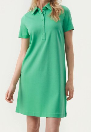 Kobieta w zielonej sukience polo z krótkim rękawem, kołnierzykiem i pięcioma guzikami, stojąca na jednolitym jasnym tle.