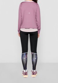 Maglia atletica a maniche lunghe mauve sopra un sotto leggero rosa, abbinata a leggings neri con un motivo astratto colorato e sneakers viola chiaro.