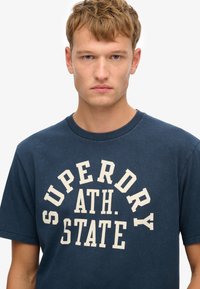 Camiseta de algodón azul marino con mangas cortas y cuello redondo, que presenta el texto en blanco "SUPERDRY ATH. STATE" en letras gruesas en el pecho.