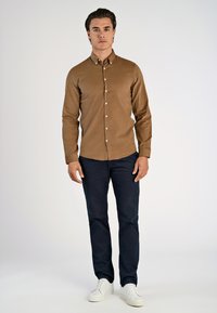 Camicia marrone a maniche lunghe con colletto button-down, abbinata a pantaloni blu scuro e sneaker bianche, in piedi contro uno sfondo chiaro.