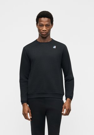 Mann som har på seg en enkel svart langermet genser med en liten fargerik logo på venstre bryst, stående foran en hvit bakgrunn.