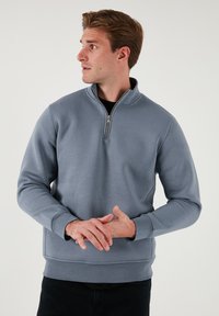 Felpa con zip di colore grigio chiaro realizzata in tessuto liscio. Presenta un colletto alto e polsini a costine. Indossata con pantaloni neri.