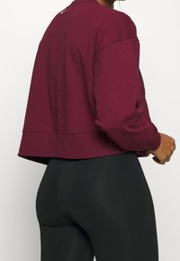 Žena nosící zkrácenou bordó mikinu a černé legíny s vysokým pasem, stojící bokem na jednobarevném světlém pozadí.