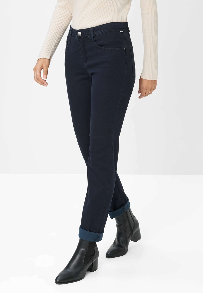 Dunkelblaue Jeans mit schmaler Passform, hohem Bund, Fronttaschen und umgeschlagenen Säumen. Kombiniert mit schwarzen Stiefeletten.