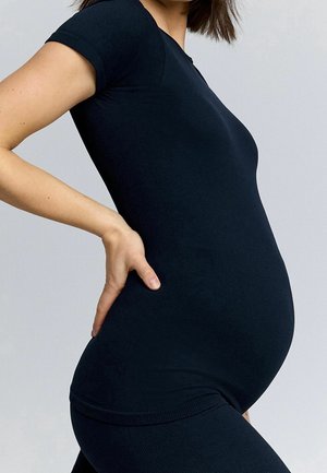 Kobieta w ciąży w czarnym obcisłym topie z krótkim rękawem i legginsach, pokazana z boku, ręka oparta na dole pleców na jednolitym tle.