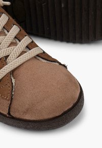 Sneaker in tela marrone con punta arrotondata, superficie testurizzata, lacci beige e suola in gomma nera. Presenta accenti di cucitura sui lati.