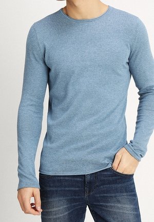 Homme portant une chemise ajustée bleu clair à manches longues et un jean bleu foncé, debout avec une main dans la poche devant un fond neutre.