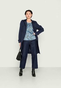 Casaco de trench azul-marinho, blusa com padrão floral, calças largas de ganga escuras, botas pretas de tornozelo e uma mala preta de ombro. Texturas suaves.
