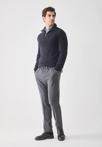 Granatowy sweter dzianinowy z kołnierzem na zamek, szare spodnie na miarę oraz czarne skórzane buty. Sweter ma teksturowany wzór.