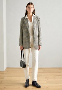 Blazer a quadri nei toni grigi, neri e crema; camicia bianca; pantaloni avorio; borsa nera testurizzata; stivali blu navy; sfondo in legno.