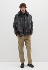 Giubbotto bomber in pelle marrone con colletto peloso, indossato sopra una camicia a quadri. Pantaloni beige e scarpe nere completano l'outfit.