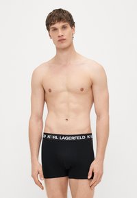 Melni kokvilnas bokseršorti ar platu melnu jostu, uz kuras baltos burtiem rakstīts zīmola nosaukums, gluda tekstūra un piegulošs dizains.