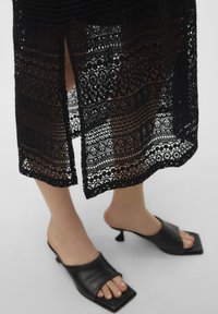 Rochie neagră din dantelă cu modele intricate și o fanta laterală, asociată cu mule negre cu partea din față pătrată și un toc mic.