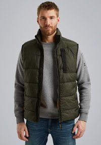 Padded olivgrüne Weste mit hohem Kragen, schwarzem Reißverschluss und Logodetail. Über einem grauen Sweatshirt getragen und mit blauen Jeans kombiniert.