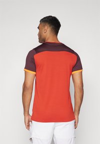 Punainen ja viininpunainen urheilupaita lyhyillä hihoilla, jossa on oranssit yksityiskohdat hihoissa ja teksturoitu, hengittävä kangasmateriaali.