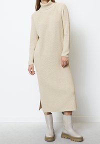 Longue robe en maille beige avec col roulé et fente latérale, portée avec des bottines à semelle épaisse beige clair, sur fond blanc.
