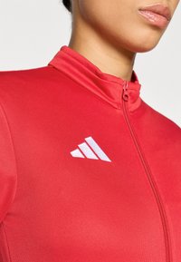 Κόκκινο αθλητικό μπουφάν με φερμουάρ στον γιακά, λείο ύφασμα, με λευκό λογότυπο Adidas στο αριστερό στήθος.