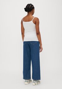 Hvit tettsittende tanktop med tynne stropper, kombinert med løse blå denimjeans. Modellen har på seg hvite joggesko, som viser et avslappet antrekk bakfra.