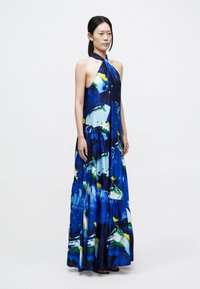 Pitkä, halterneck-mallinen mekko virtaavasta kankaasta. Siinä on sininen abstrakti kuvio vihreillä ja keltaisilla yksityiskohdilla. Mekossa on leveä helma, jossa on kerroksittainen leikkaus.