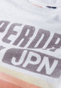 Camiseta de algodón gris claro con una gran impresión desvanecida que dice "SUPERDRY JPN" en gris oscuro. Presenta un escote redondo y una textura suave.