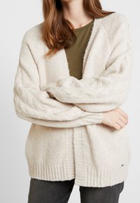 Světle béžová pletená kardigán s žebrovaným lemem a rukávy s kabelkovým vzorem, má volnější střih a otevřený přední design. Nositelný přes tmavě zelený top.