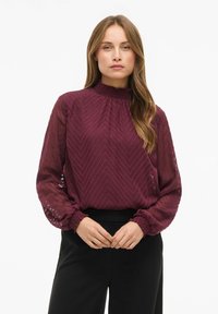 Mujer con cabello largo y castaño claro, vistiendo una blusa burdeos transparente con textura en chevron y pantalones negros, de pie frente a un fondo blanco.