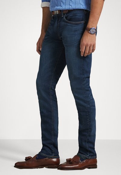 Σκούρο μπλε τζιν με στενή εφαρμογή, φτιαγμένο από τζιν, με αντίθετες ραφές και υποτυπώδη υφή. Συνδυάζεται με καφέ loafers και ρολόι.