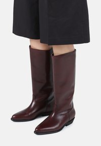 Bottes en cuir bordeaux montantes jusqu'au genou avec une texture lisse et un bout pointu, dotées d'un petit talon carré et de simples accents de couture.