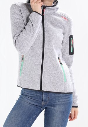 Person trägt eine hellgraue Zip-up-Jacke mit grün akzentuierten Reißverschlusstaschen und blaue Jeans, Hand nahe am Gesicht.