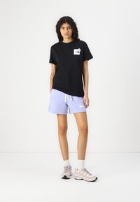 Črna bombažna majica s kratkimi rokavi in žepom na prsih, v kombinaciji s svetlo vijoličnimi kratkimi hlačami z elastičnim pasom in logotipom.