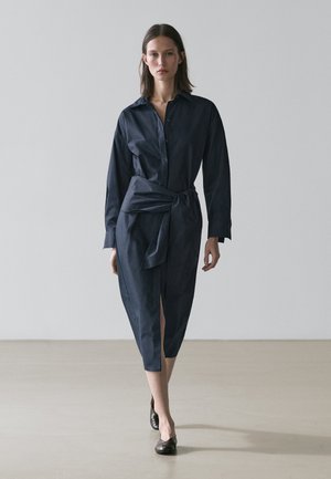 Kobieta idąca w ciemnoniebieskiej, długorękawowej sukience zapinanej na guziki, z przednim rozcięciem i dużym paskiem w talii, nosząca czarne baleriny.