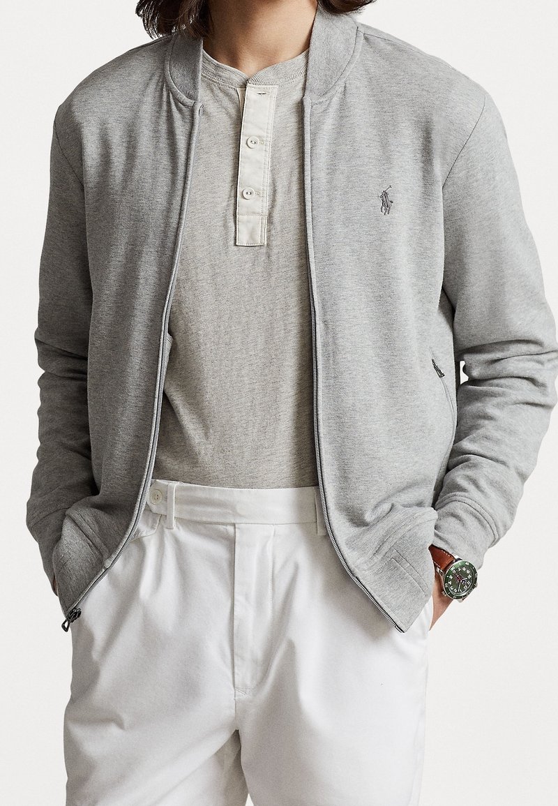 Homme portant une veste zippée gris clair par-dessus un t-shirt henley gris, un pantalon blanc et une montre verte au poignet gauche.