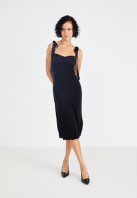 Μαύρο αμάνικο φόρεμα με εφαρμοστό μπούστο, σατέν φιόγγους στους ιμάντες και gathered κρόσσι, συνδυασμένο με μαύρες μυτερές γόβες.