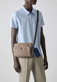 Borsa a tracolla marrone con monogramma e hardware dorato, con due scomparti con zip e un'etichetta blu. Indossata con una polo azzurra chiara e pantaloni beige.