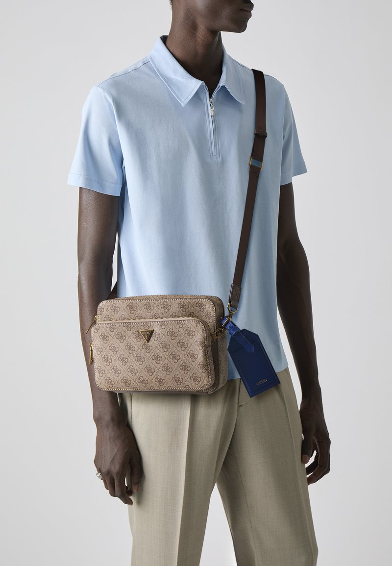 Borsa a tracolla marrone con monogramma e hardware dorato, con due scomparti con zip e un'etichetta blu. Indossata con una polo azzurra chiara e pantaloni beige.