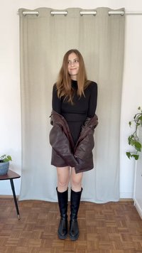 Jaqueta de couro castanha sobre um vestido preto justo, combinada com botas pretas até ao joelho. O fundo apresenta uma cortina clara e uma planta em vaso.