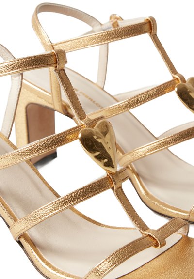 Sandali alti con cinturini dorati caratterizzati da cinturini metallici texturizzati, un accento a forma di cuore e una soletta beige liscia.