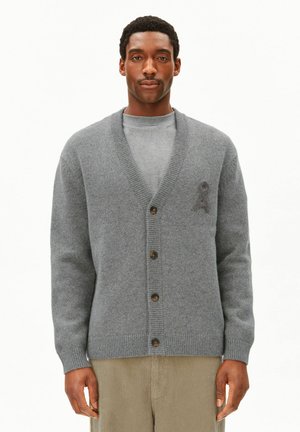 Mann trägt eine graue, geknöpfte Strickjacke über einem grauen Hemd und beigen Hosen, steht vor einem schlichten weißen Hintergrund.