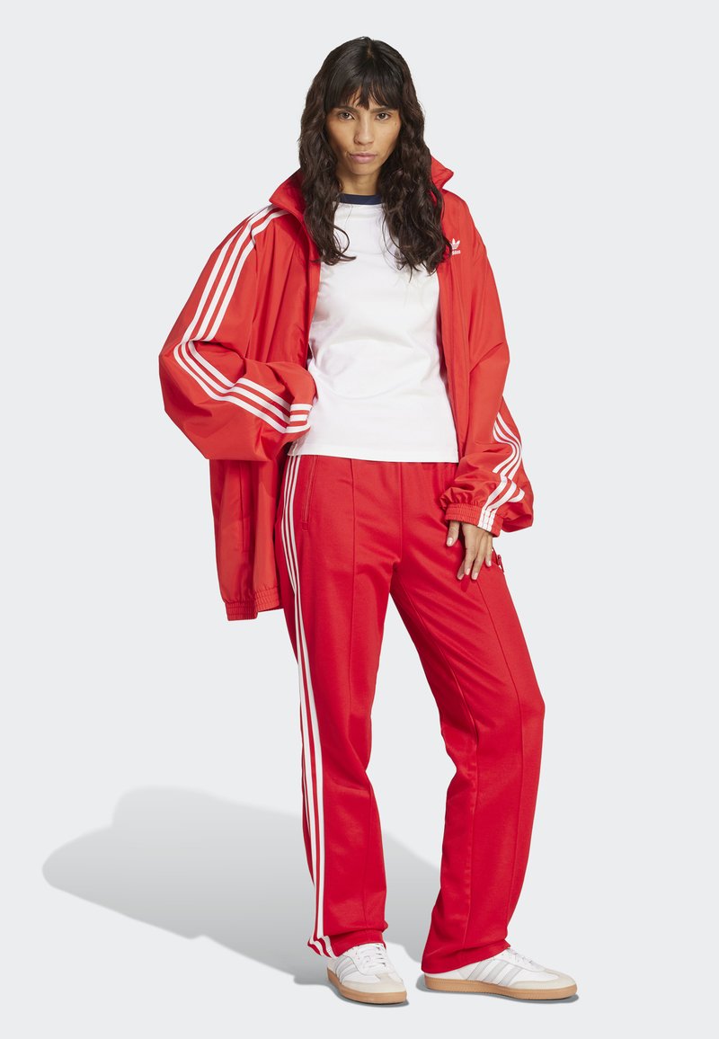 Röd idrottsjacka med vita ränder, tillsammans med matchande röda byxor. Outfiten inkluderar en vit T-shirt och vita sneakers.