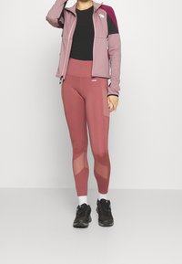 Sportjacka i ljuslila och mörk burgunder, ihop med åtsittande mörkrosa leggings med nätpaneler. Svarta sneakers fulländar looken.