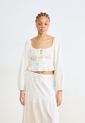 White two-piece outfit featuring a smocked crop top with long sleeves and a matching tiered skirt with a drawstring waist.