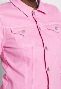 Cazadora vaquera de color rosa claro con cuello, dos bolsillos en el pecho y botones de tono plateado. Presenta detalles de costuras en la parte frontal.