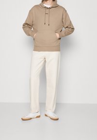 Sudadera beige con bolsillo frontal y capucha ajustable, combinada con pantalones color crema y zapatillas blancas con suelas marrones.