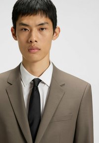 Light brown suit jacket over a white dress shirt, complemented by a black tie. The jacket features a single chest pocket and structured lapels.