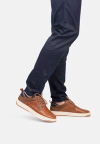 Zapatillas de cuero marrón con detalles perforados, talón negro en contraste y suela blanca. Combinadas con pantalones azul marino, mostrando un look casual.