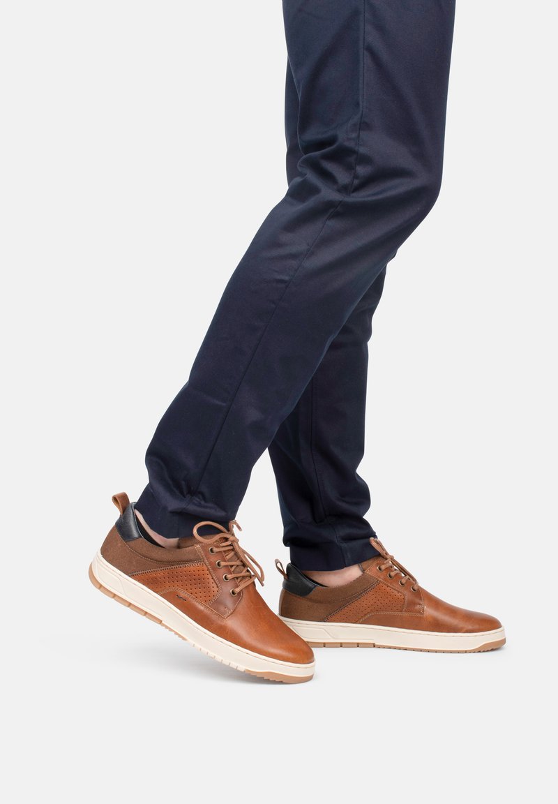 Zapatillas de cuero marrón con detalles perforados, talón negro en contraste y suela blanca. Combinadas con pantalones azul marino, mostrando un look casual.