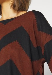 Top de punto con un patrón en zigzag en rojo oscuro y negro, con un escote amplio y una textura suave, exhibido sobre un fondo liso.