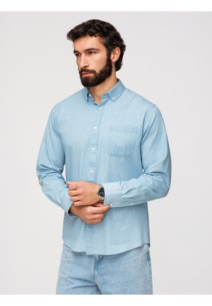 Mand med mørkt hår og skæg iført en lyseblå mønstret skjorte med knapper, lyse jeans og et armbåndsur, stående foran en ensfarvet baggrund.