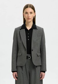 Šedý kostkovaný blazer s černým límcem, dvěma předními kapsami a zapínáním na jeden knoflík, v kombinaci s černým polo tričkem a širokými kalhotami.